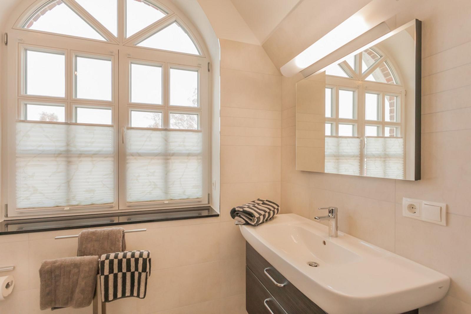 Badezimmer mit Waschbecken, Spiegel und großem Fenster mit Jalousien.