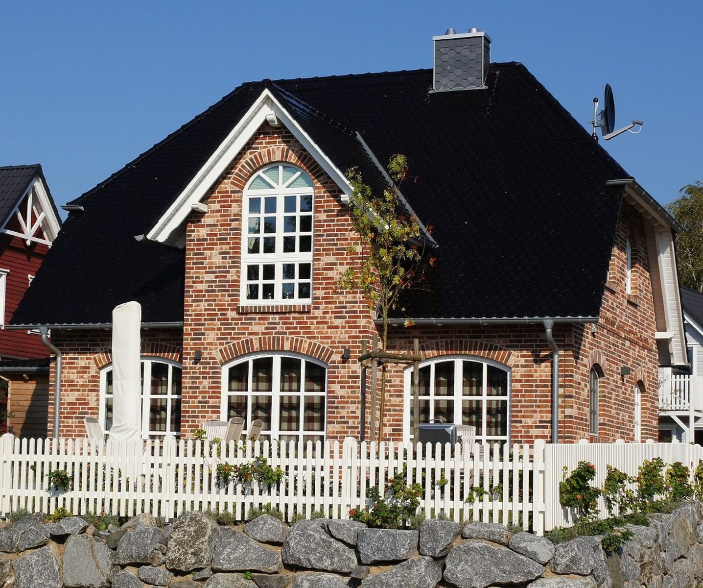 Rotes Backsteinhaus mit schwarzem Dach und weißem Zaun. Garten mit Steinen und Pflanzen.
