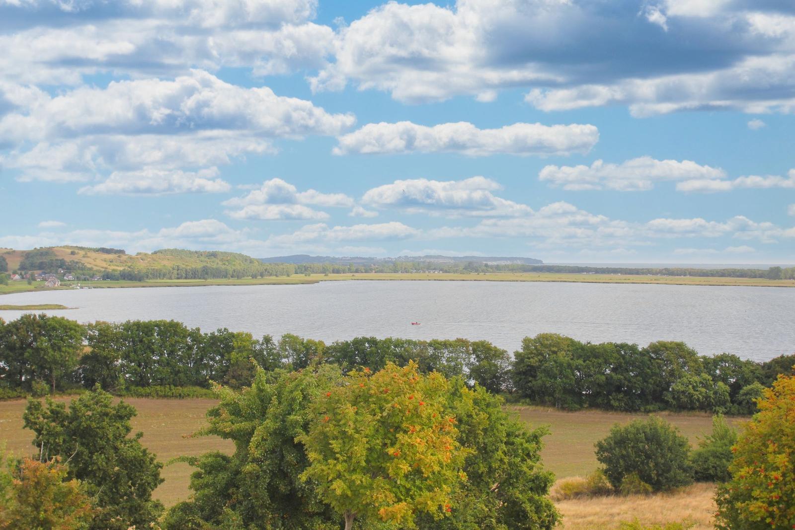 Weitläufiger See mit kleinem Boot, umgeben von Wiesen und Bäumen unter blauem Himmel mit weißen Wolken.