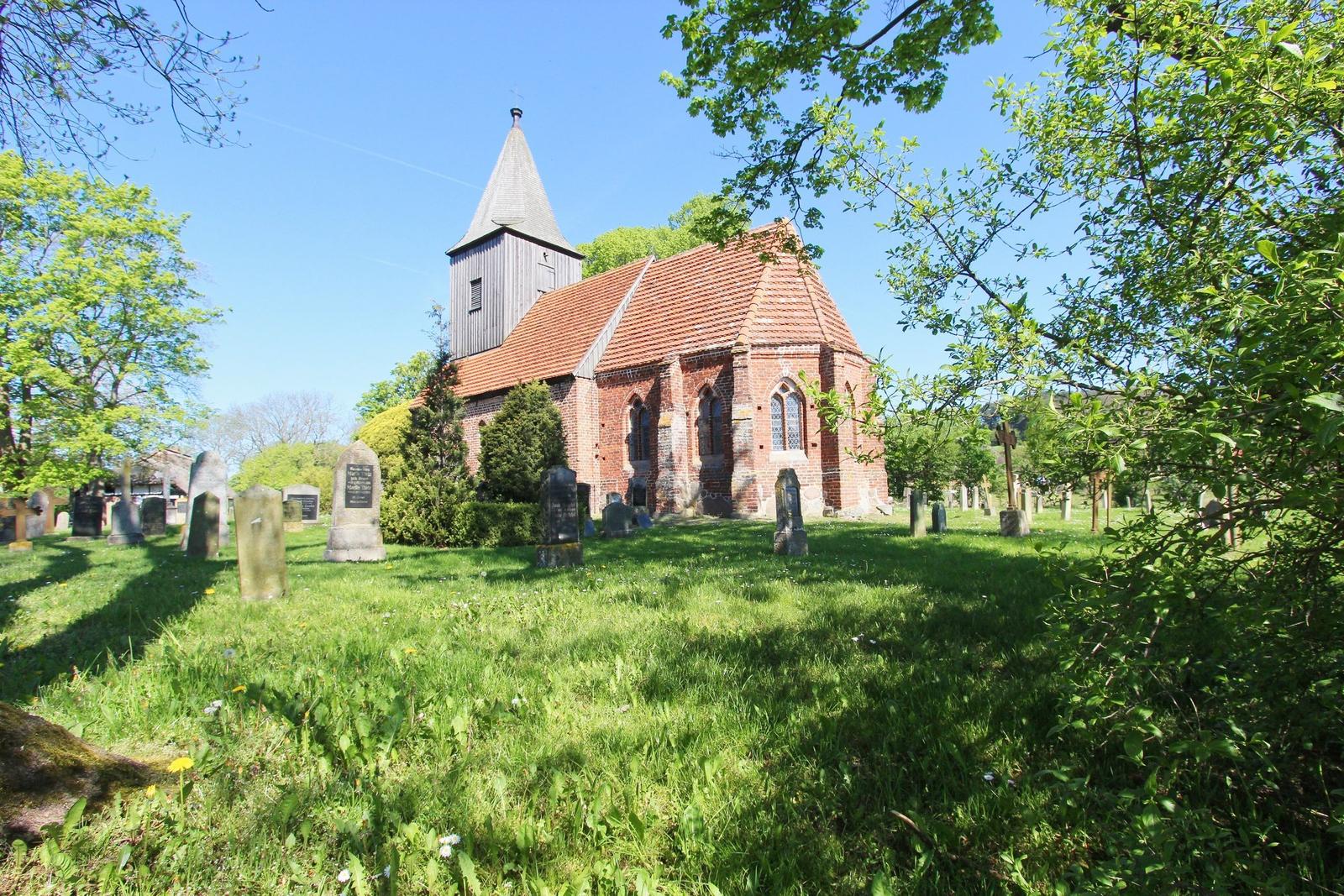 Roter Backsteinbau mit Turm und Friedhof. Gras und Bäume umgeben das Gebäude.
