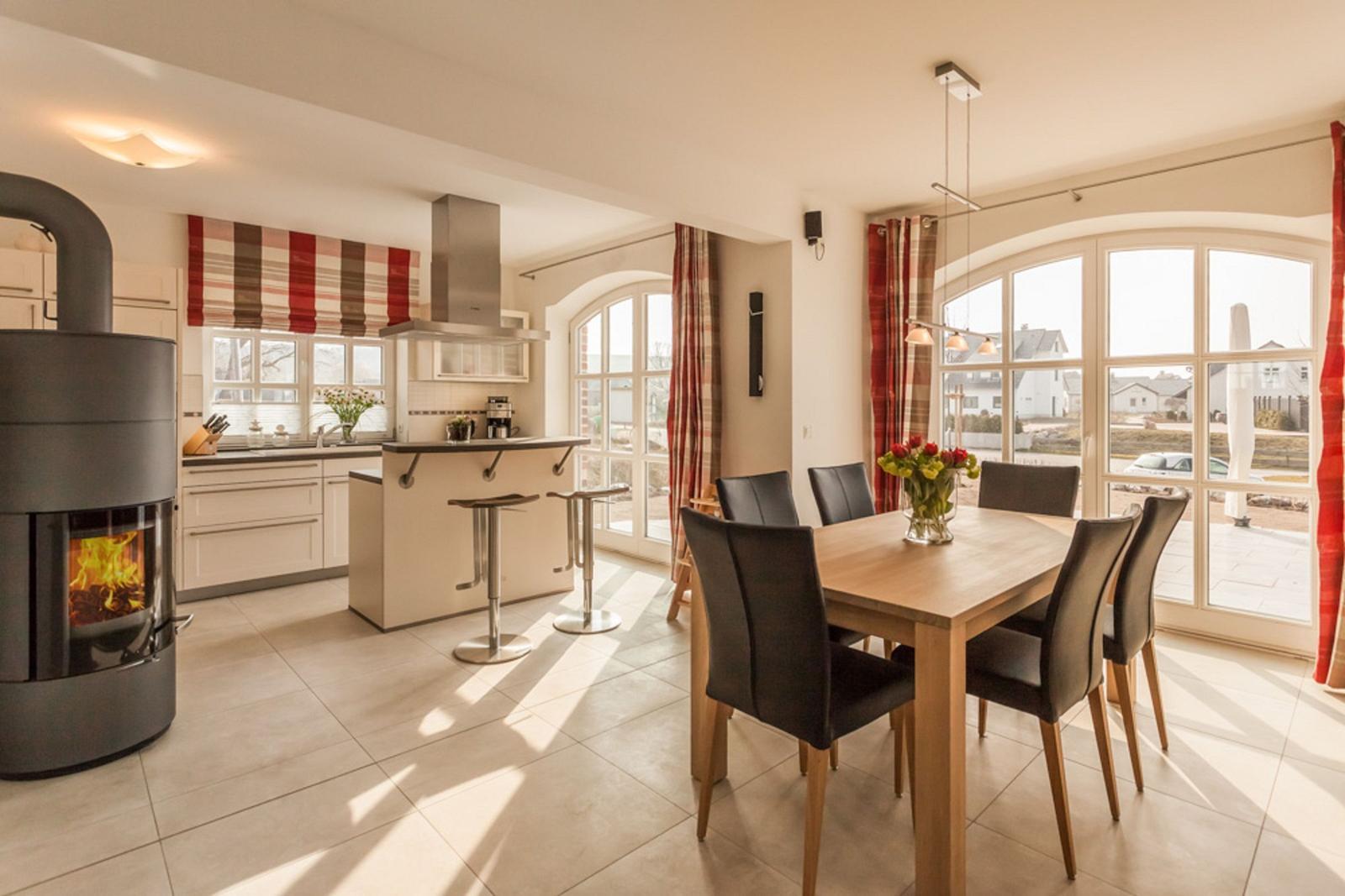 Küche und Essbereich mit Holztisch, Stühlen und Kaminofen. Große Fenster mit Blick nach draußen.
