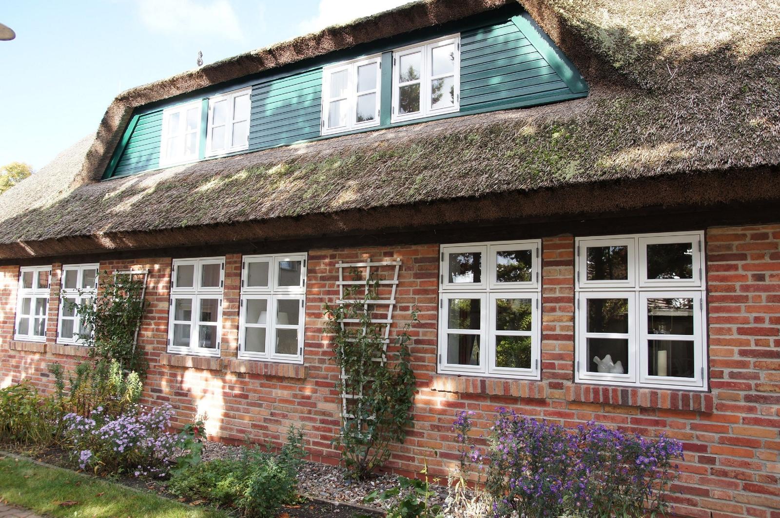 Haus mit strohgedecktem Dach und roten Ziegelmauern, weiße Fenster und Blumenbeete.