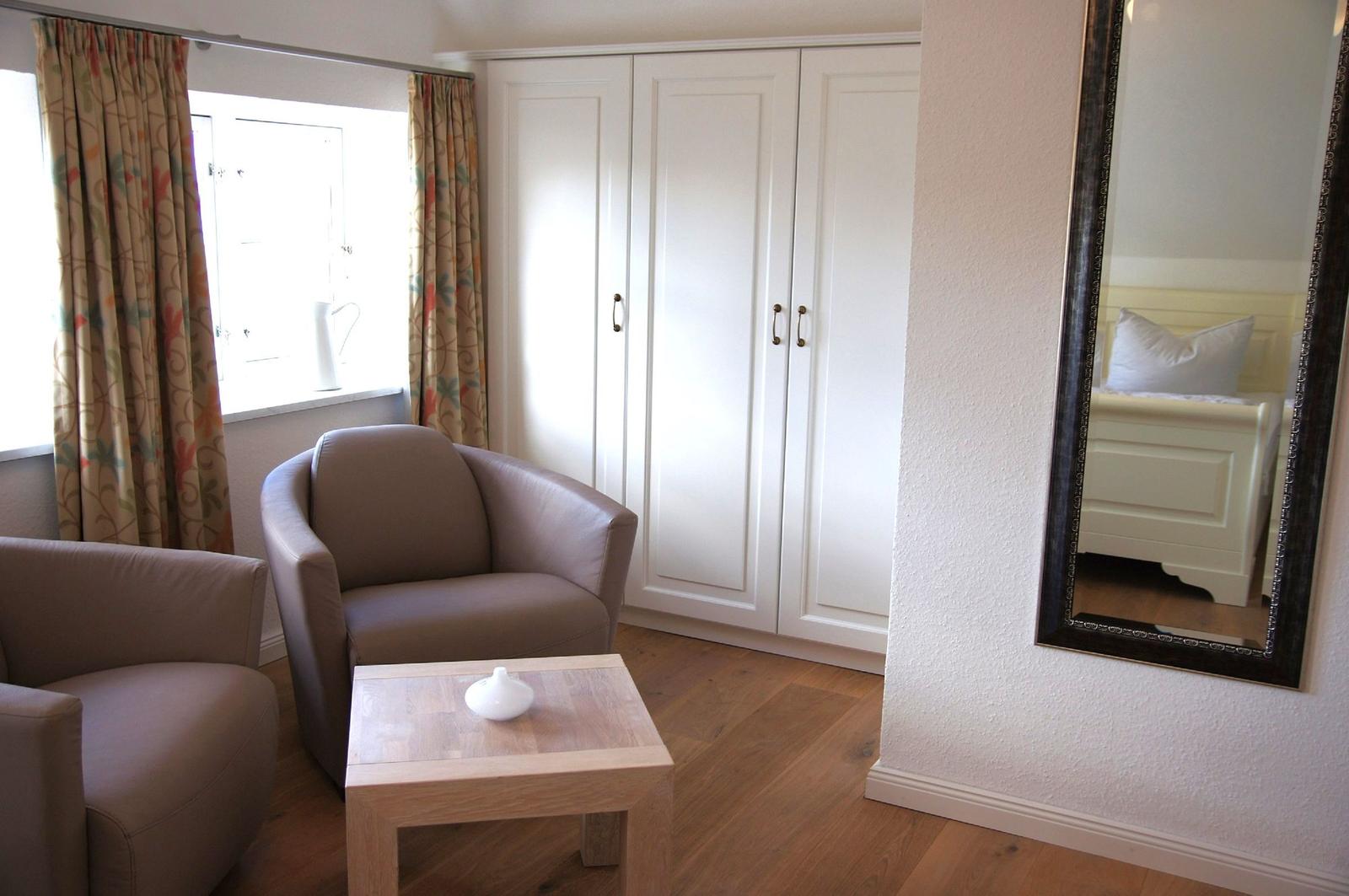 Zimmer mit Sitzgelegenheit, Kleiderschrank und Spiegel. Fenster mit Vorhängen.
