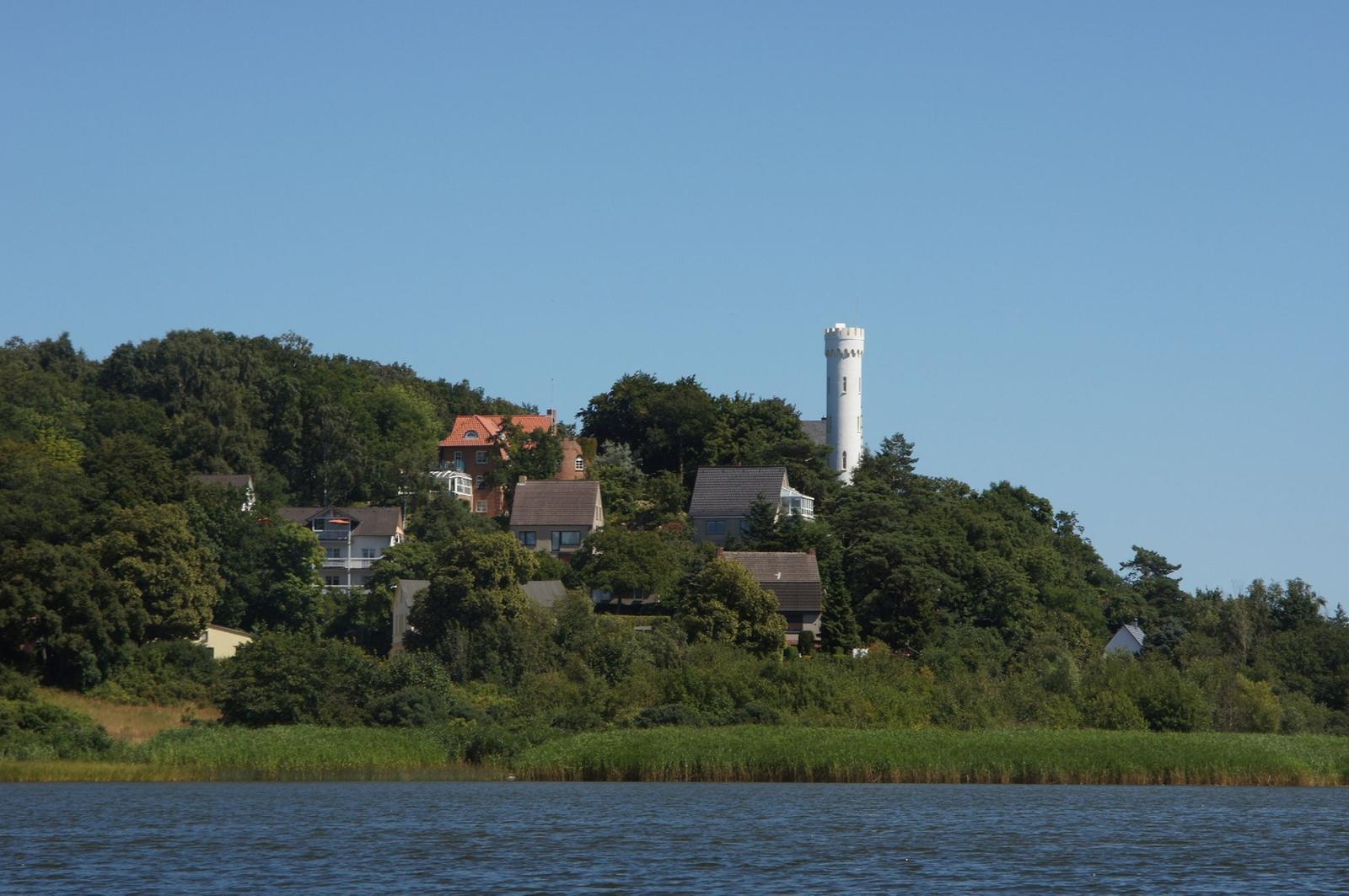 Wohnhäuser und ein weißer Turm auf einer bewaldeten Insel am See.