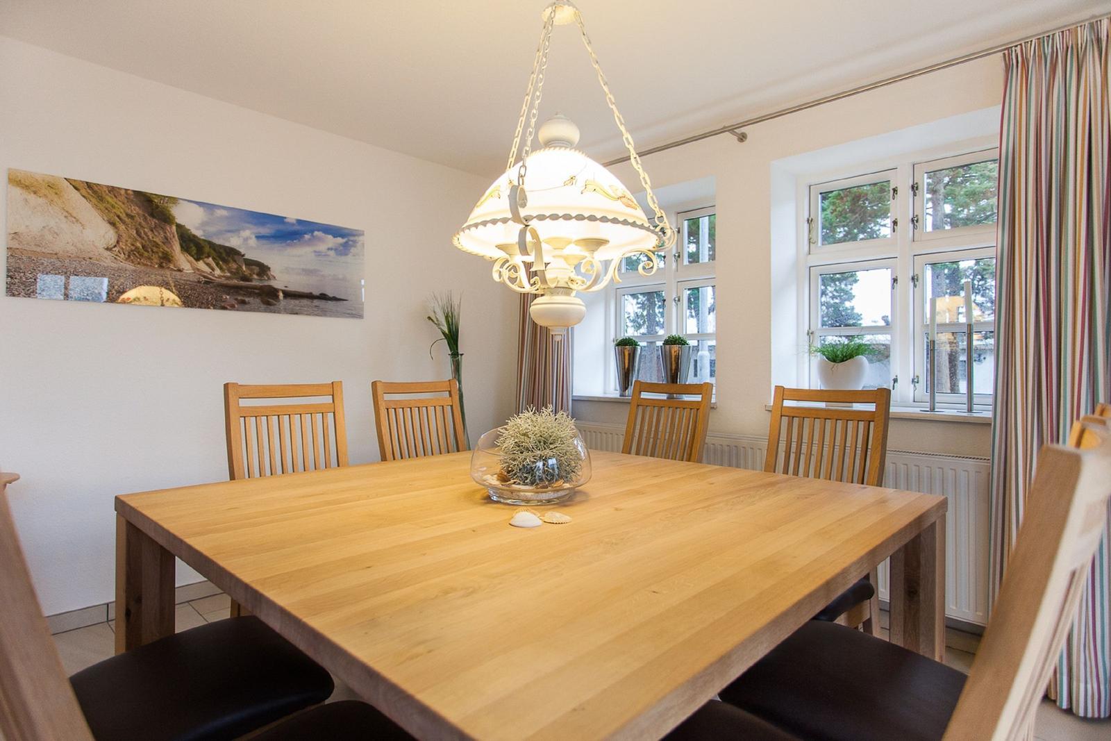 Esszimmer mit Holztisch, Stühlen und Fenster. Deckenleuchte und Wandbild sichtbar.
