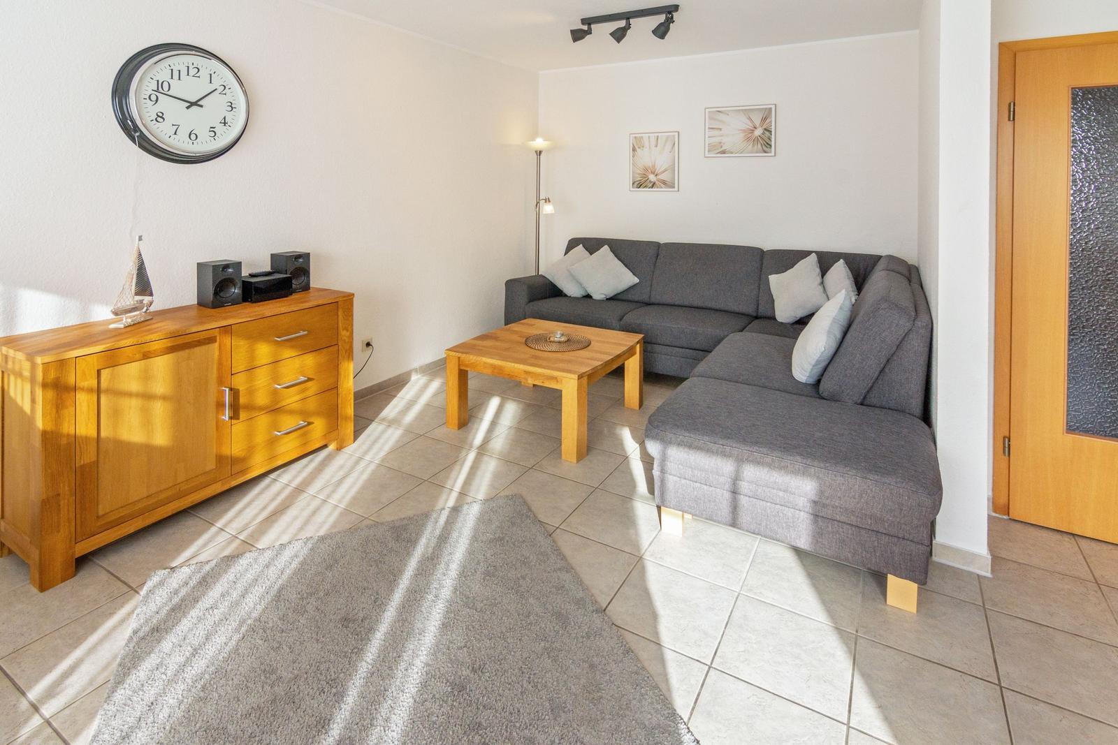 Wohnzimmer mit grauem Sofa, Holztisch und Sideboard. Wanduhr und Bilder an der Wand.