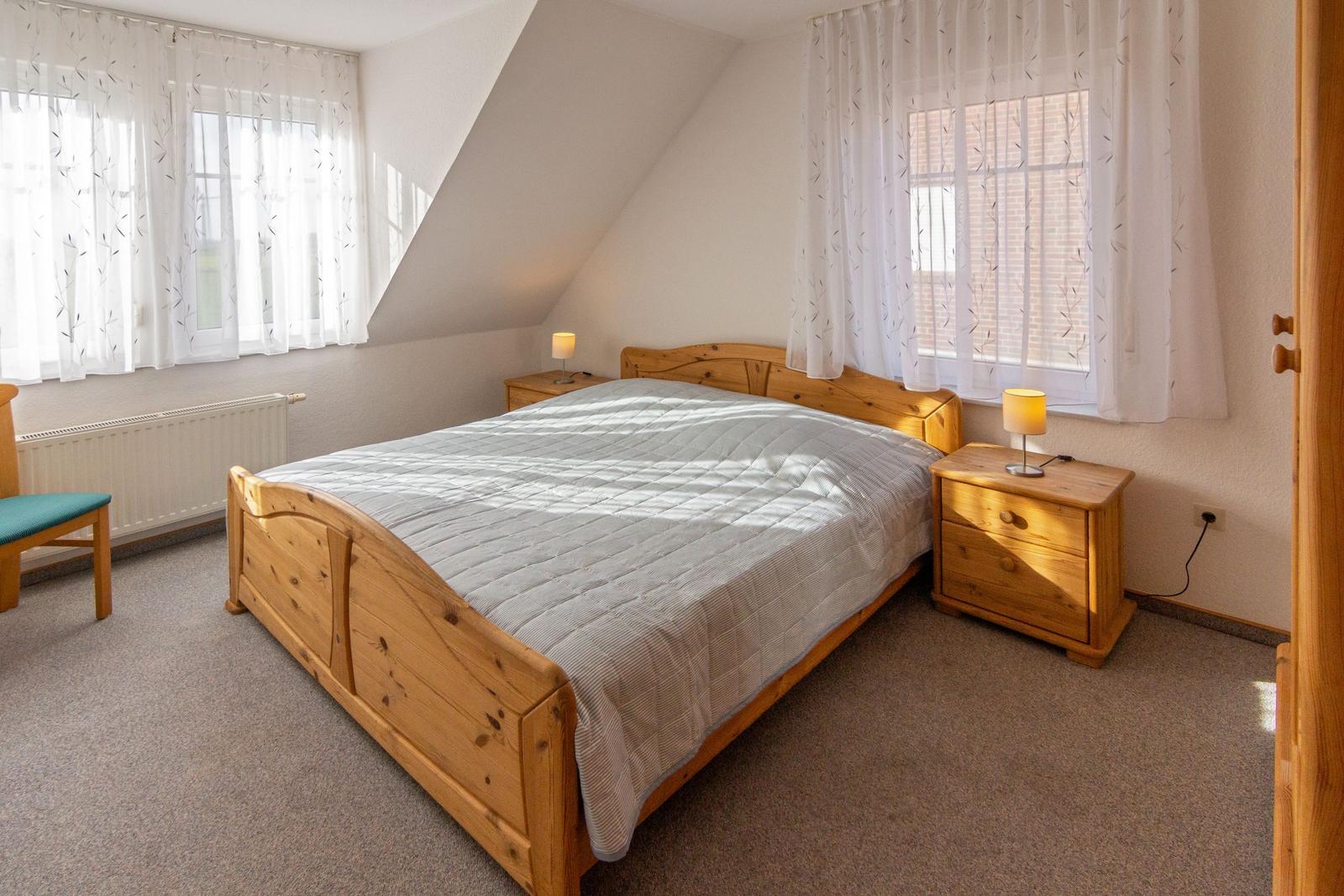 Schlafzimmer mit Holzbett, zwei Nachttischen und Fenstern mit weißen Vorhängen.