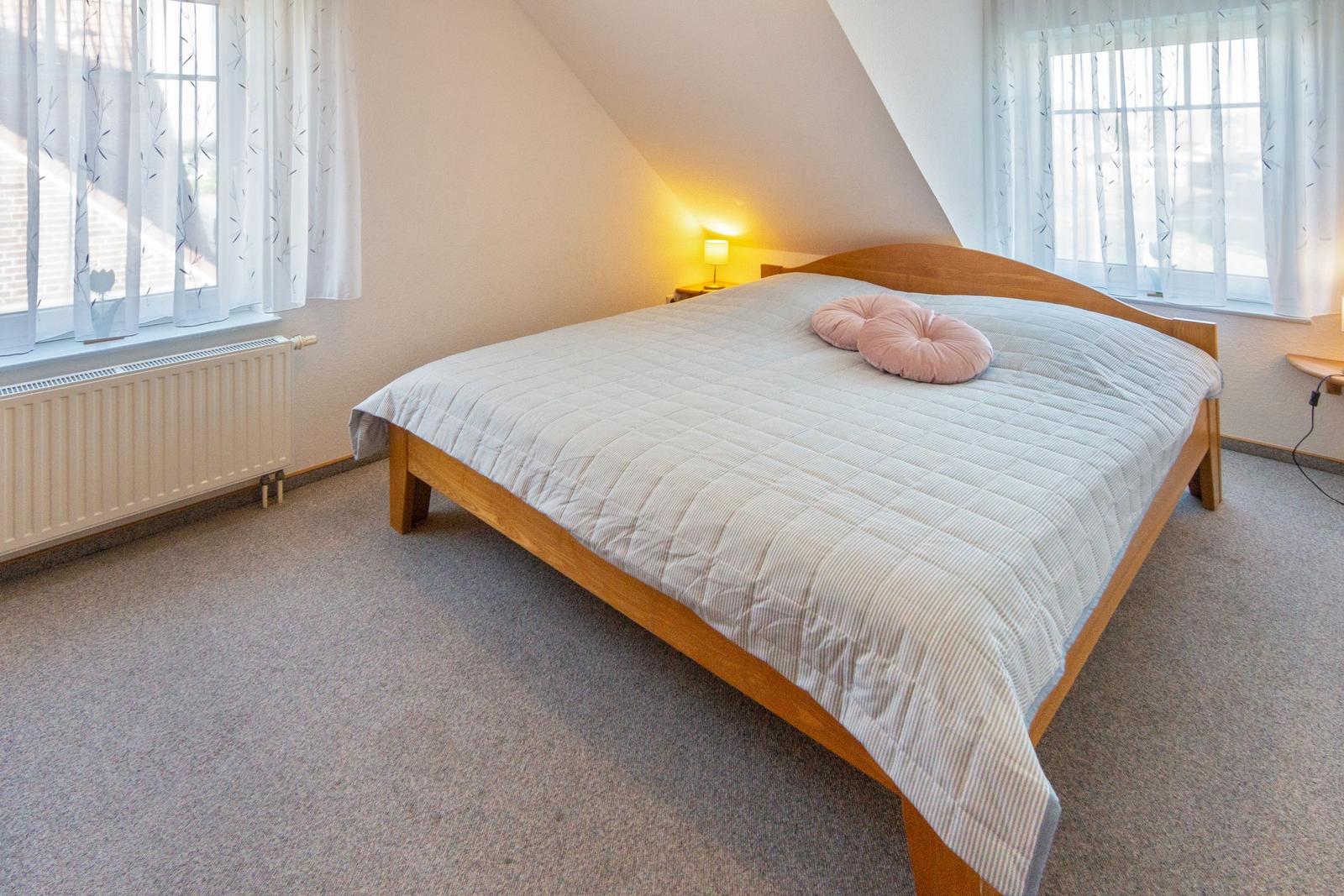 Schlafzimmer mit Holzbett, zwei Fenstern und Heizkörper an der Wand.