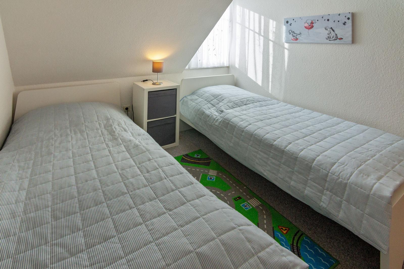 Zwei Einzelbetten, Nachttisch mit Lampe, Fenster und Kinderbild an der Wand.