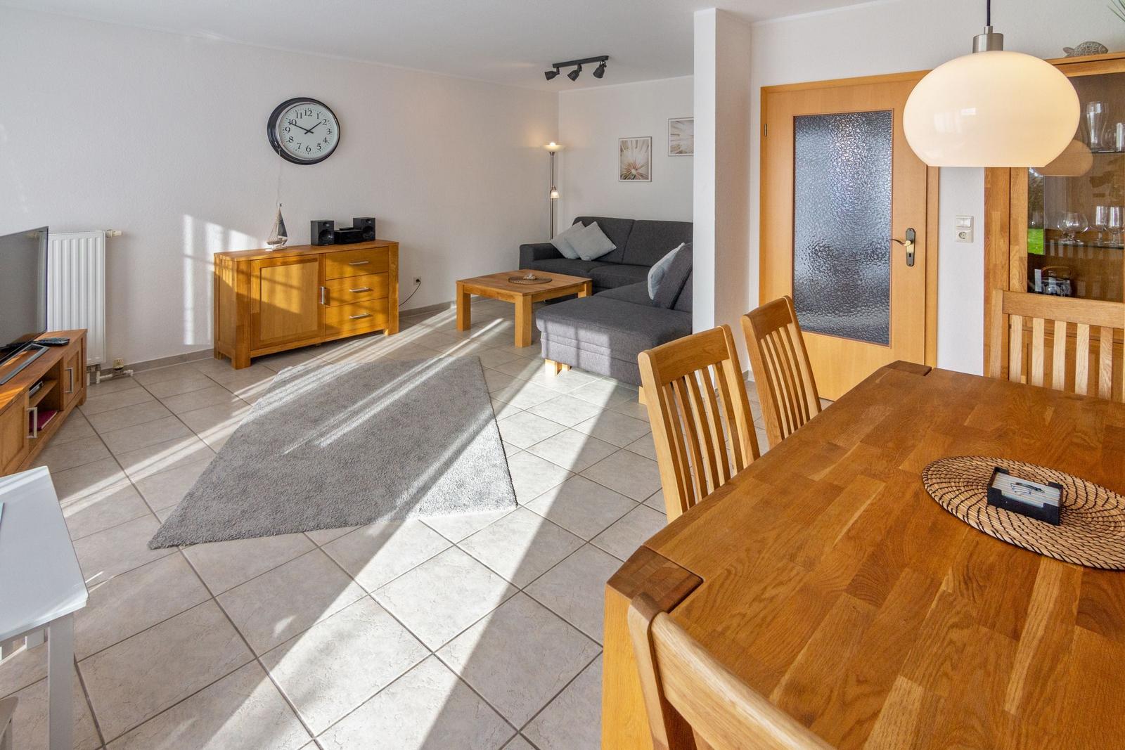 Wohn- und Essbereich mit Holzmöbeln und grauem Sofa. Sonnenlicht fällt durch Fenster.