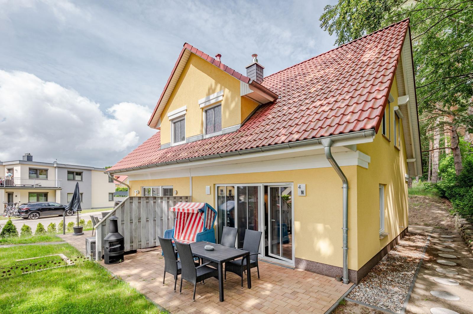 Gelbes Haus mit Terrasse, Grill und Strandkorb. Roter Ziegeldach. Grün umgeben.