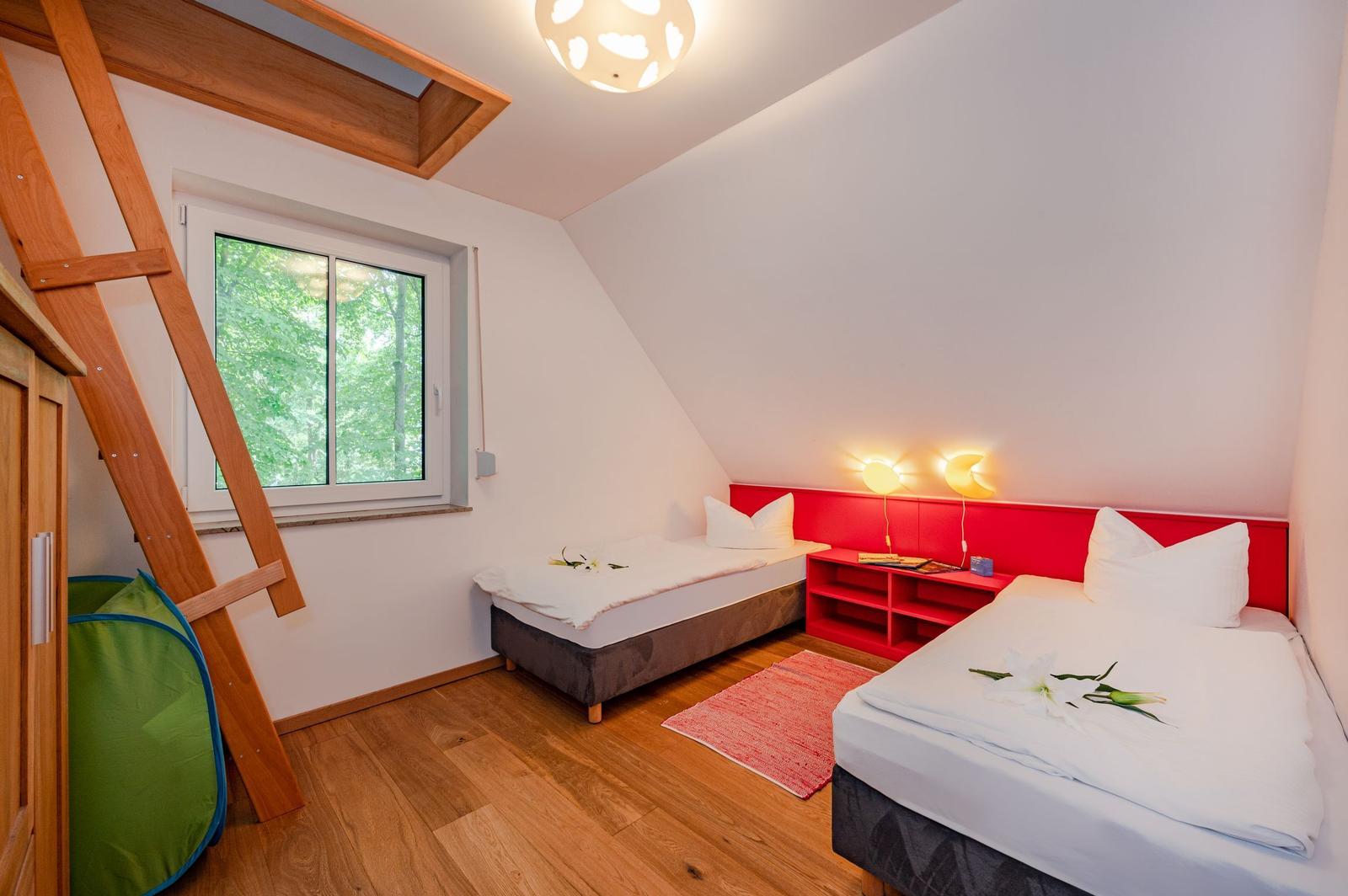 Zimmer mit zwei Betten, rotem Kopfteil und Holzboden. Fenster mit Blick auf Wald.
