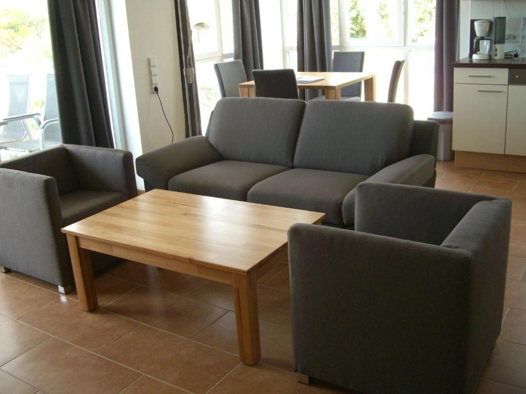 Wohnzimmer mit Sofa, Sessel und Holztisch. Küche und Essbereich im Hintergrund.