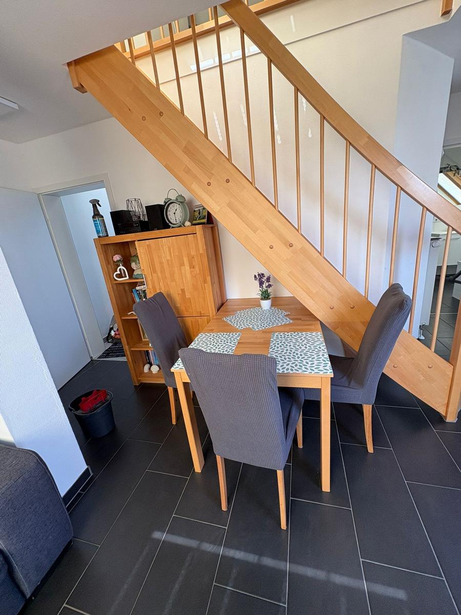 Essbereich mit Tisch, Stühlen und Holztreppe. Helle Wände und dunkler Boden.