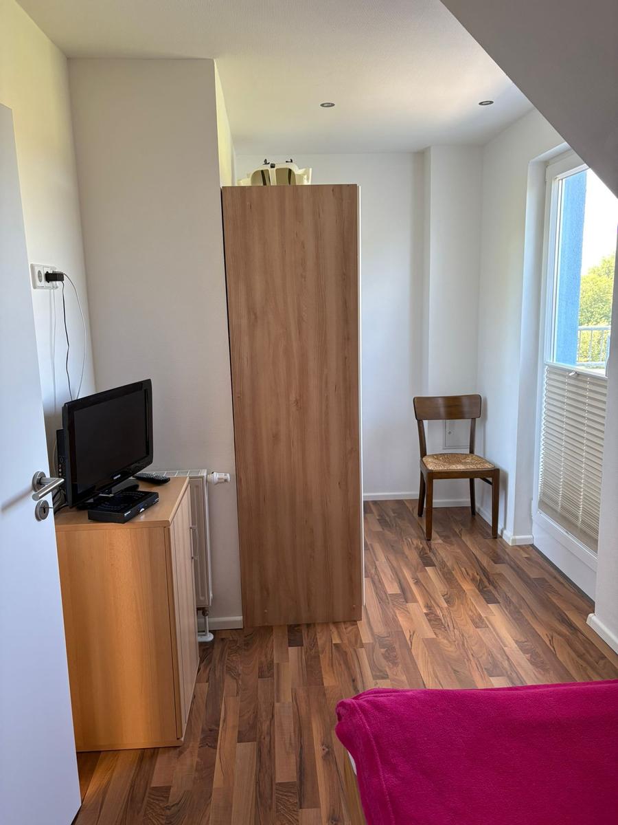 Zimmer mit Holzboden, Kleiderschrank, Stuhl und TV-Schrank. Fenster mit Jalousie.