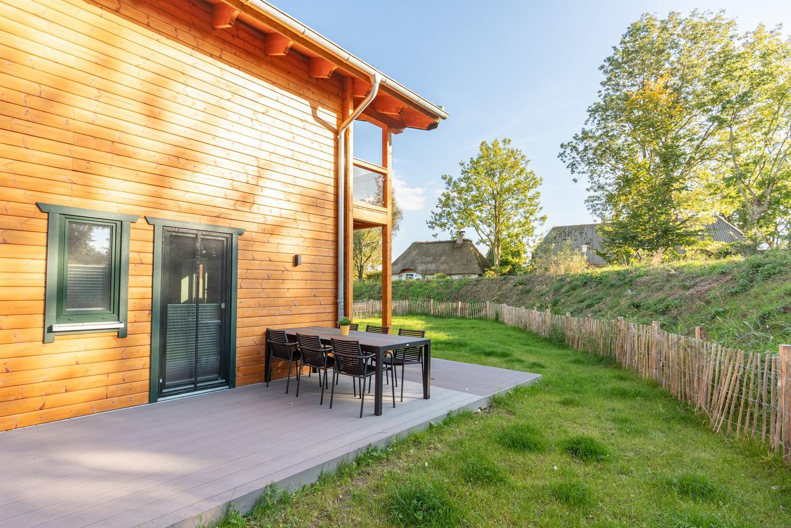 Haus mit Holzfassade und Terrasse mit Tisch und Stühlen. Grünfläche und Zaun im Hintergrund.