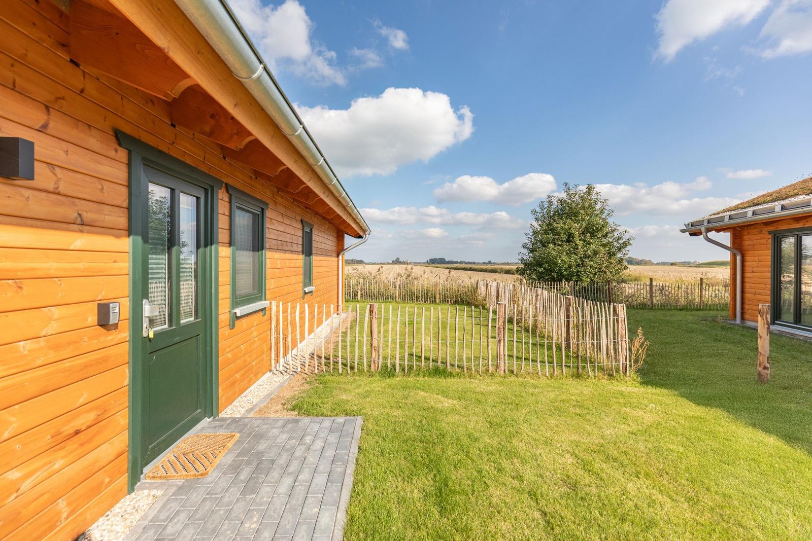 Holzhaus mit grüner Tür und Terrasse. Grasfläche und Zaun im Hintergrund.