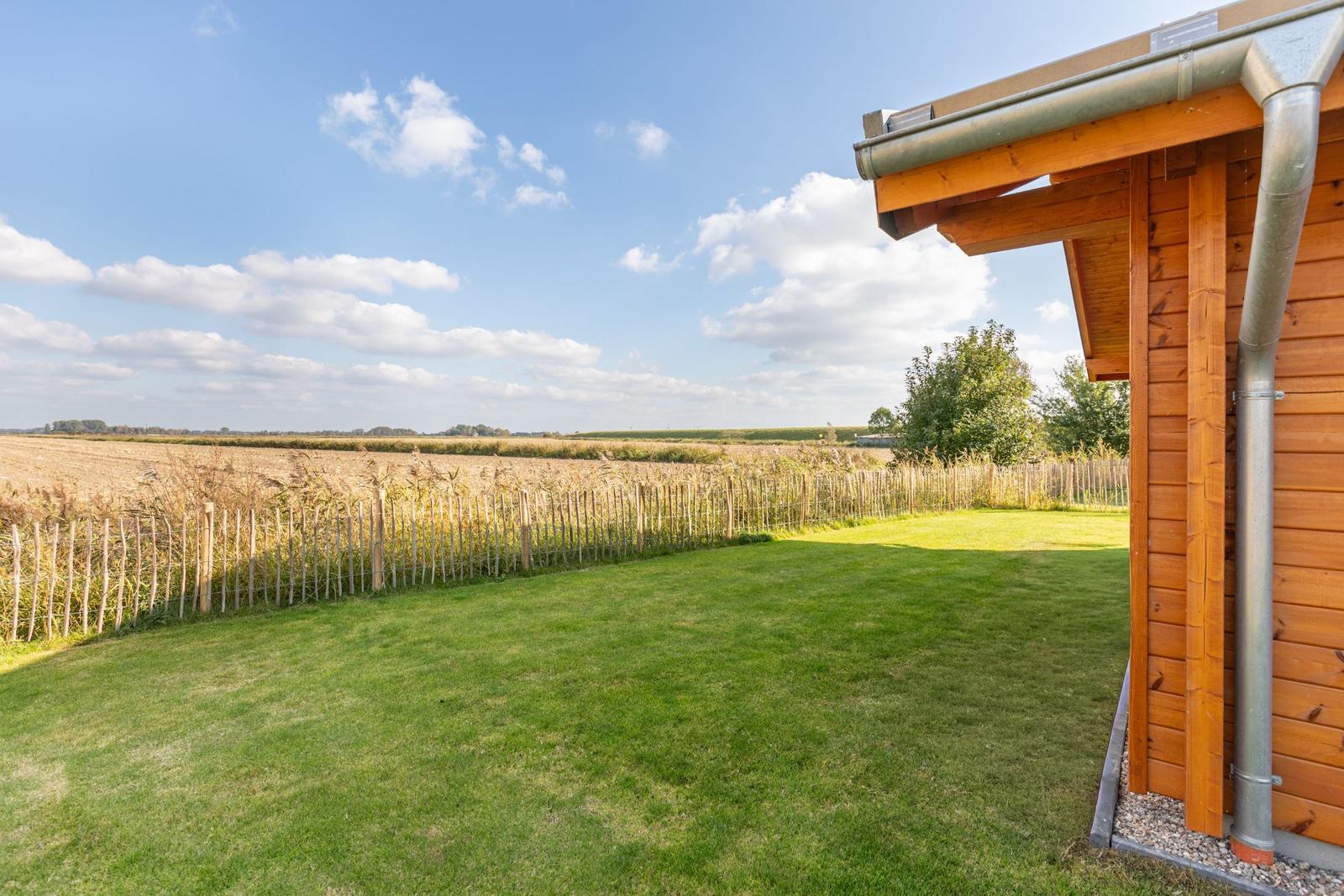 Hinter dem Holzhaus erstreckt sich ein grüner Rasenbereich mit einem Zaun und Blick auf Felder.