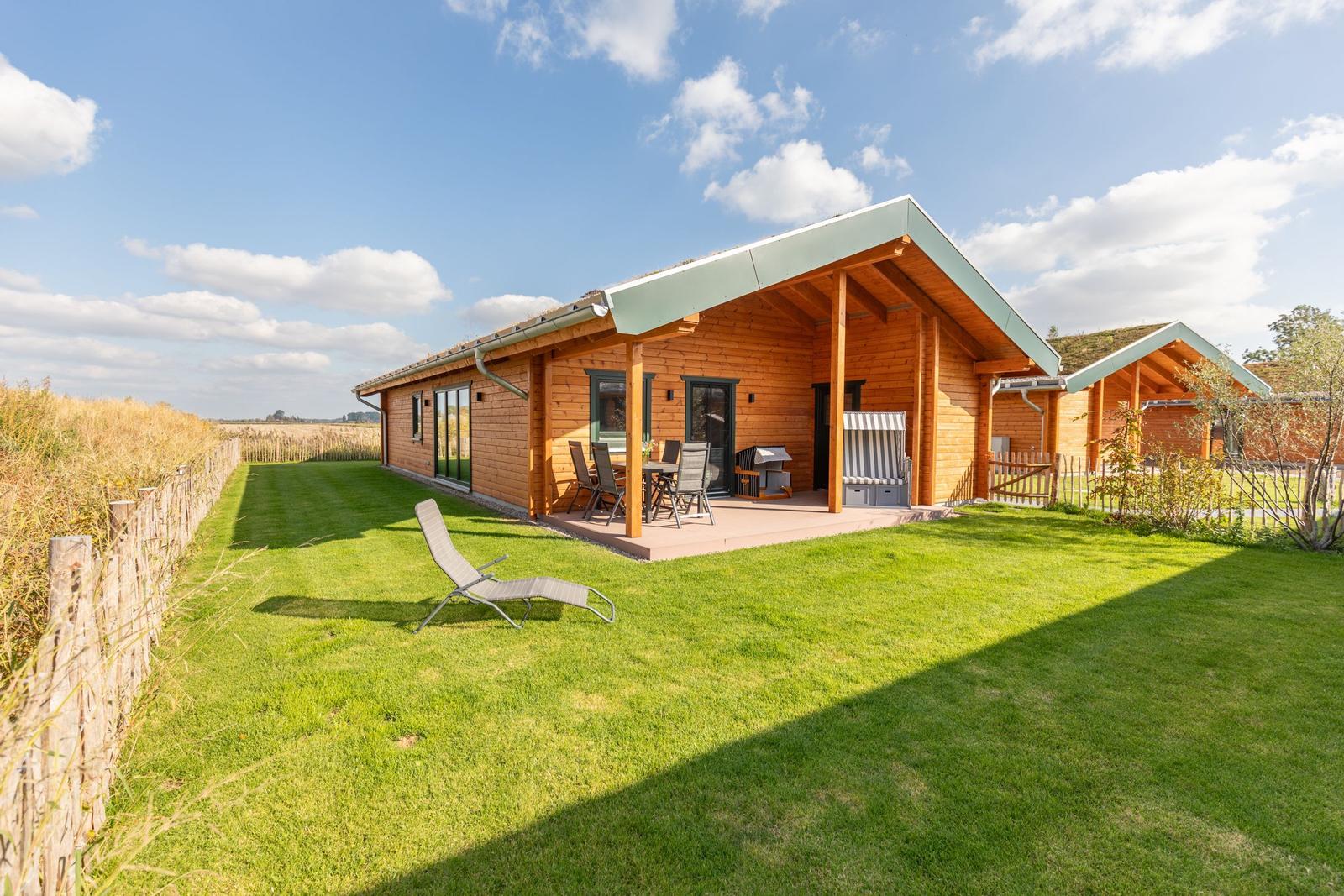 Holzhaus mit Terrasse, Garten und Liegestuhl unter blauem Himmel.