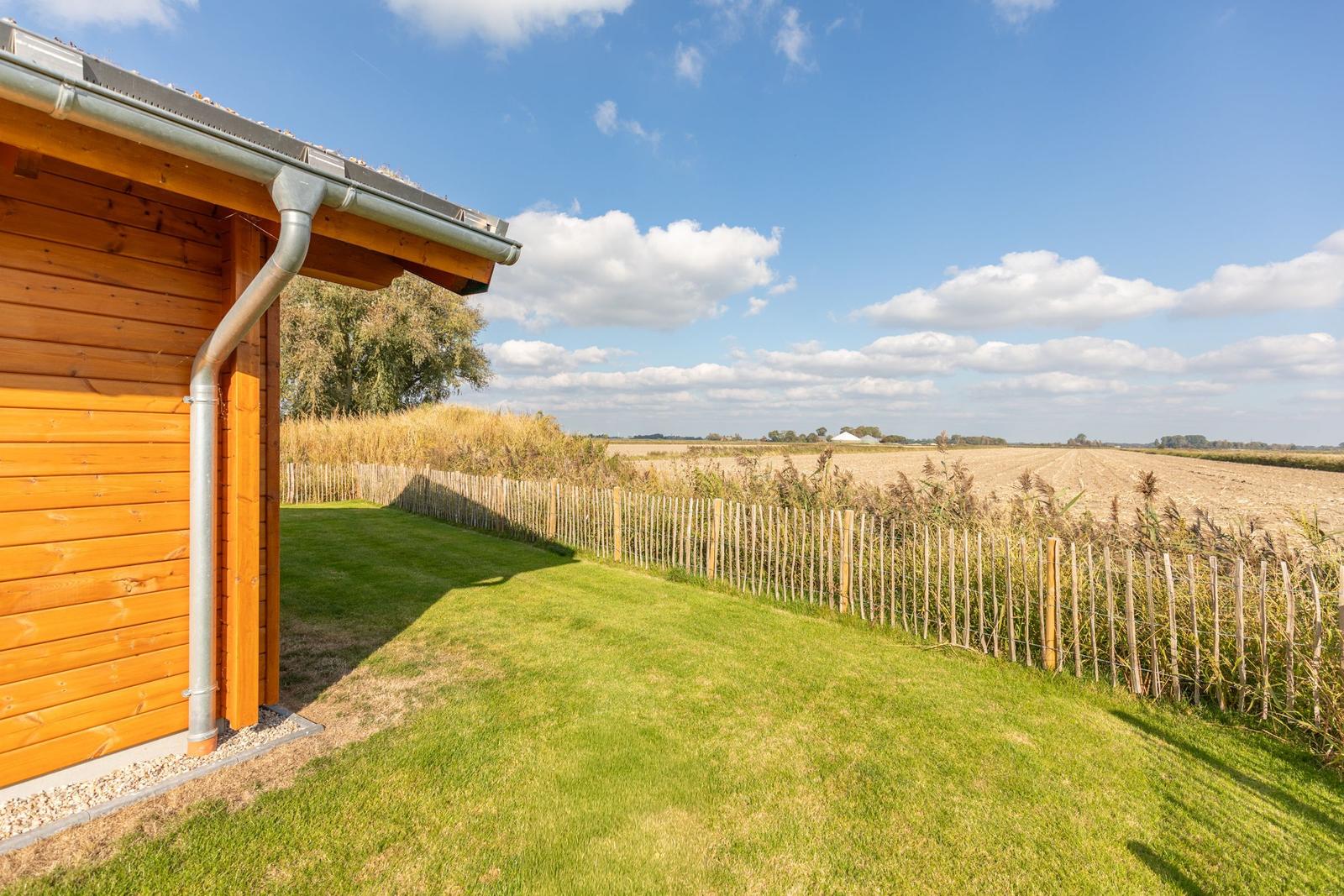 Haus mit Holzfassade, grünem Rasen und Blick auf Felder unter blauem Himmel.