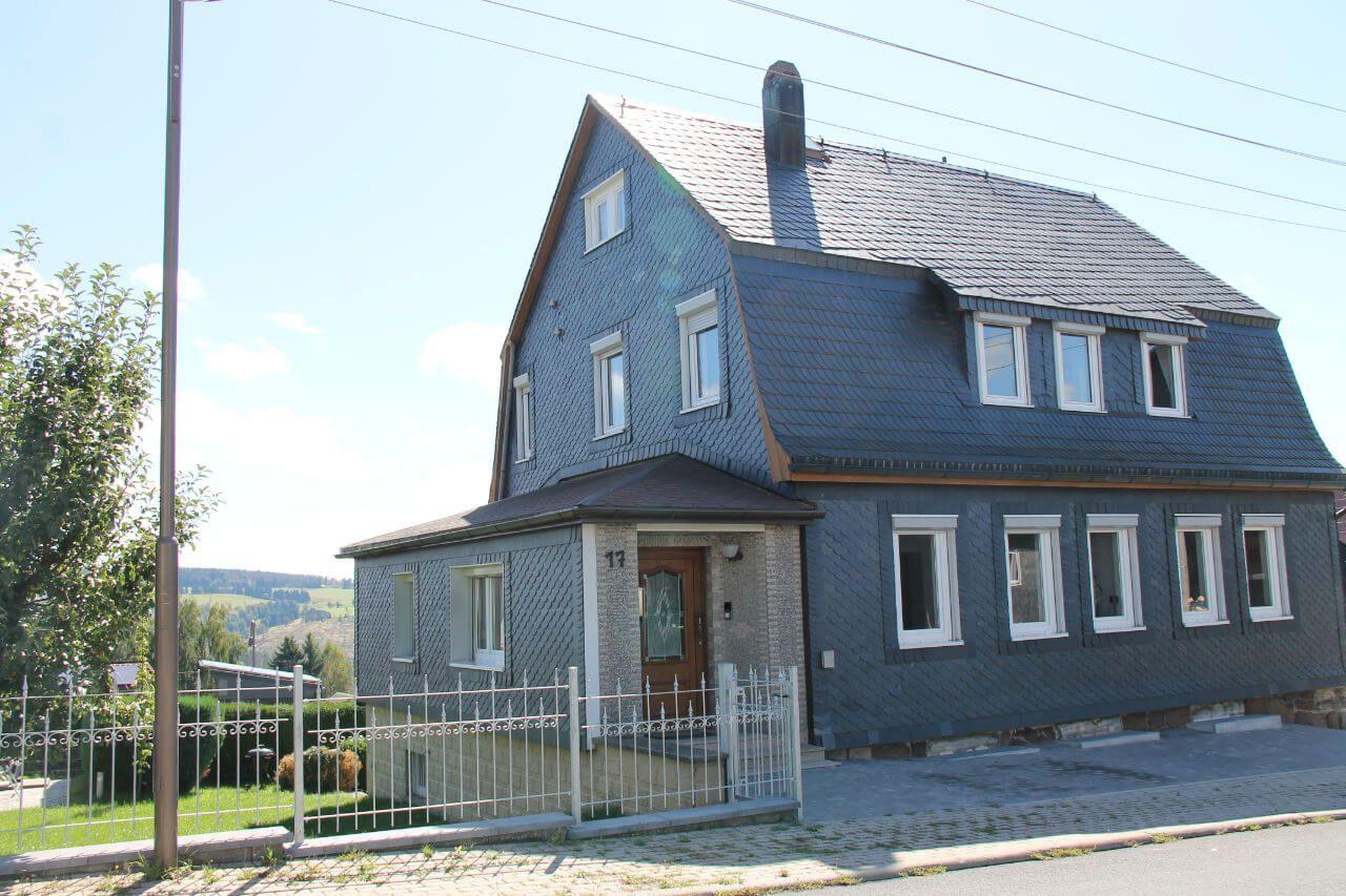 Haus mit dunklem Schieferdach und weißen Fenstern an einer Straße mit Gartenzaun.