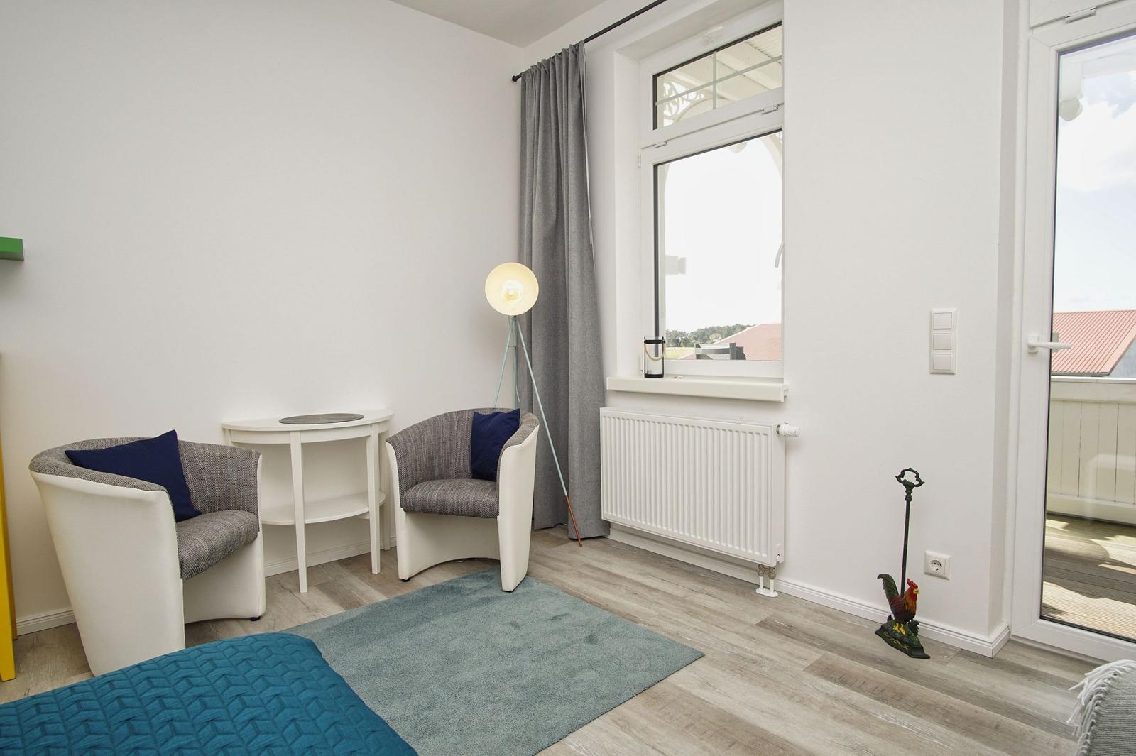 Zimmer mit zwei Sesseln, Tisch, Stehlampe und Blick auf Balkon.