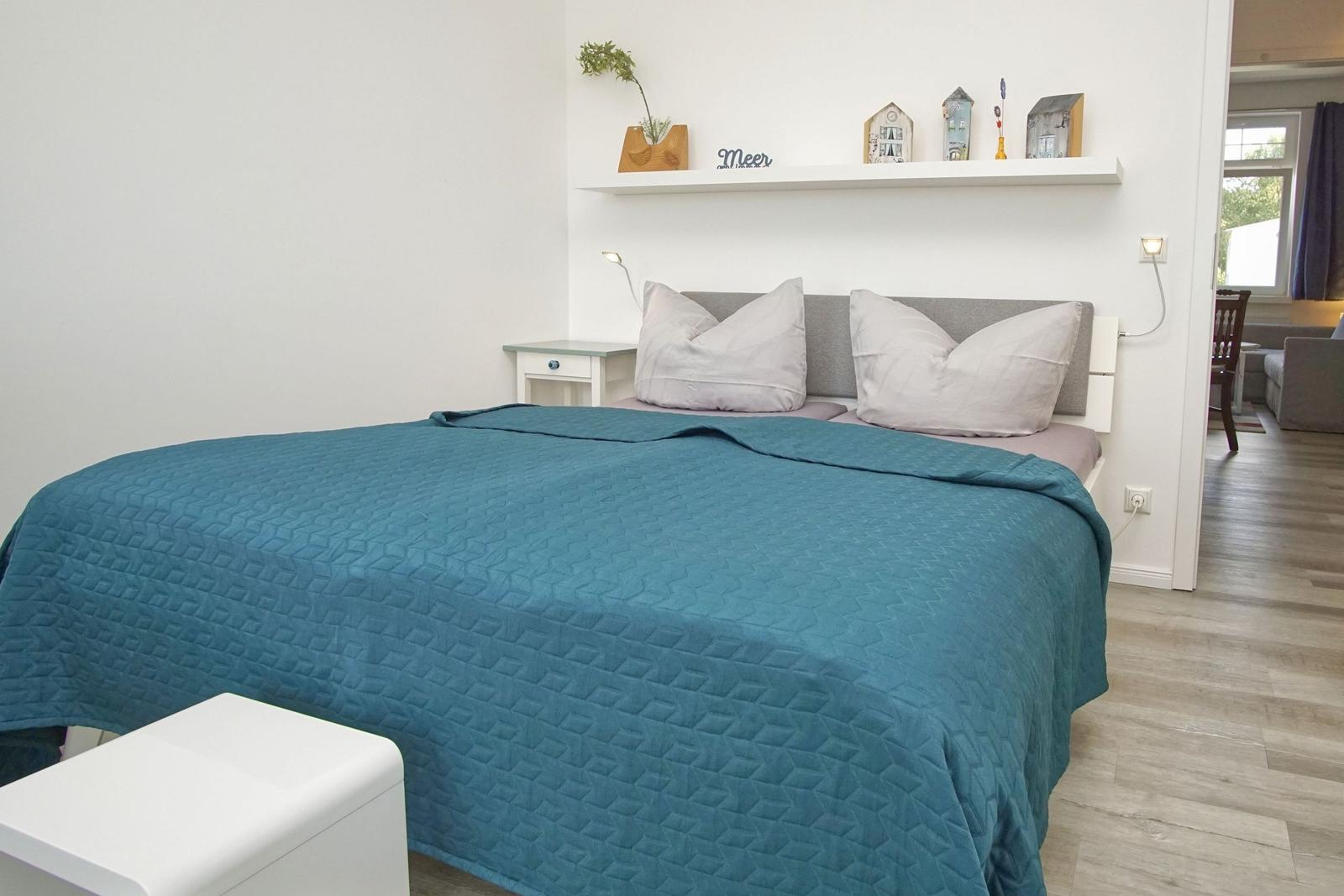 Schlafzimmer mit Bett, Nachttisch und Wandregal. Durchgang zur Wohnfläche.