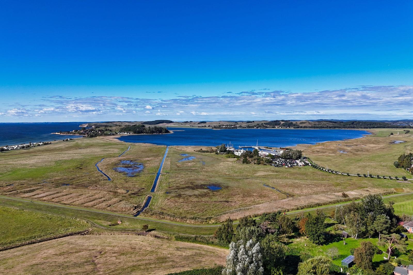 Aerielansicht eines Sees mit Uferbereichen und Grünflächen unter blauem Himmel.