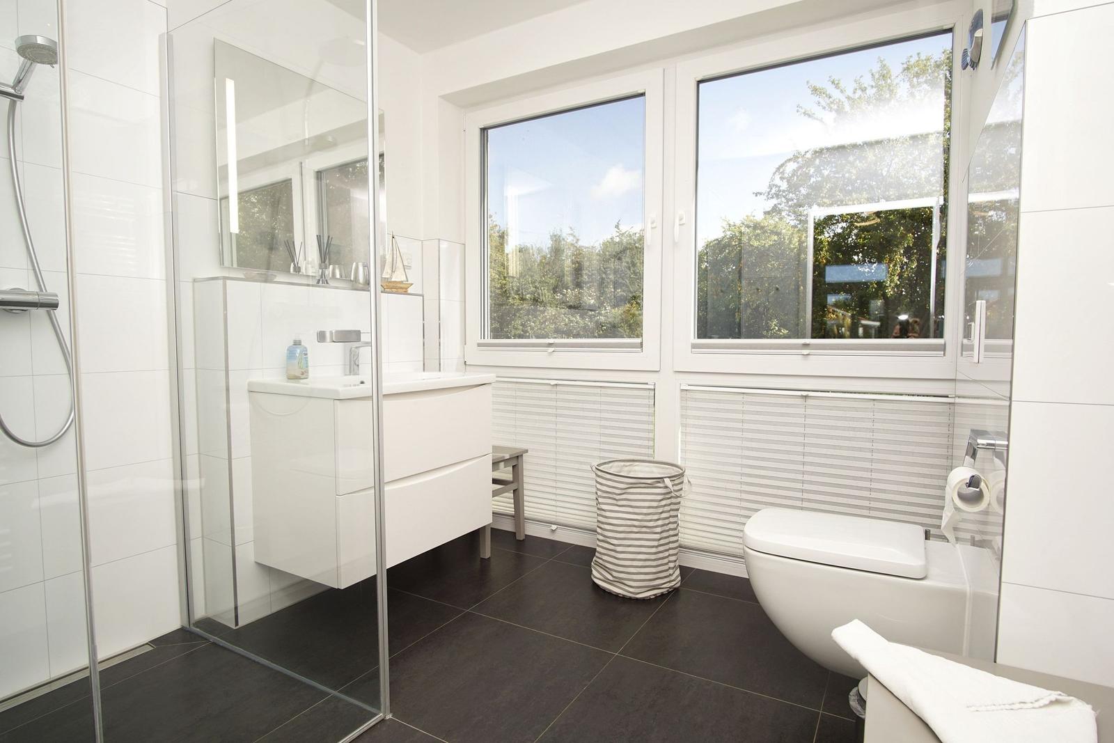 Badezimmer mit Dusche, Waschbecken und Toilette. Große Fenster mit Blick auf Grün.