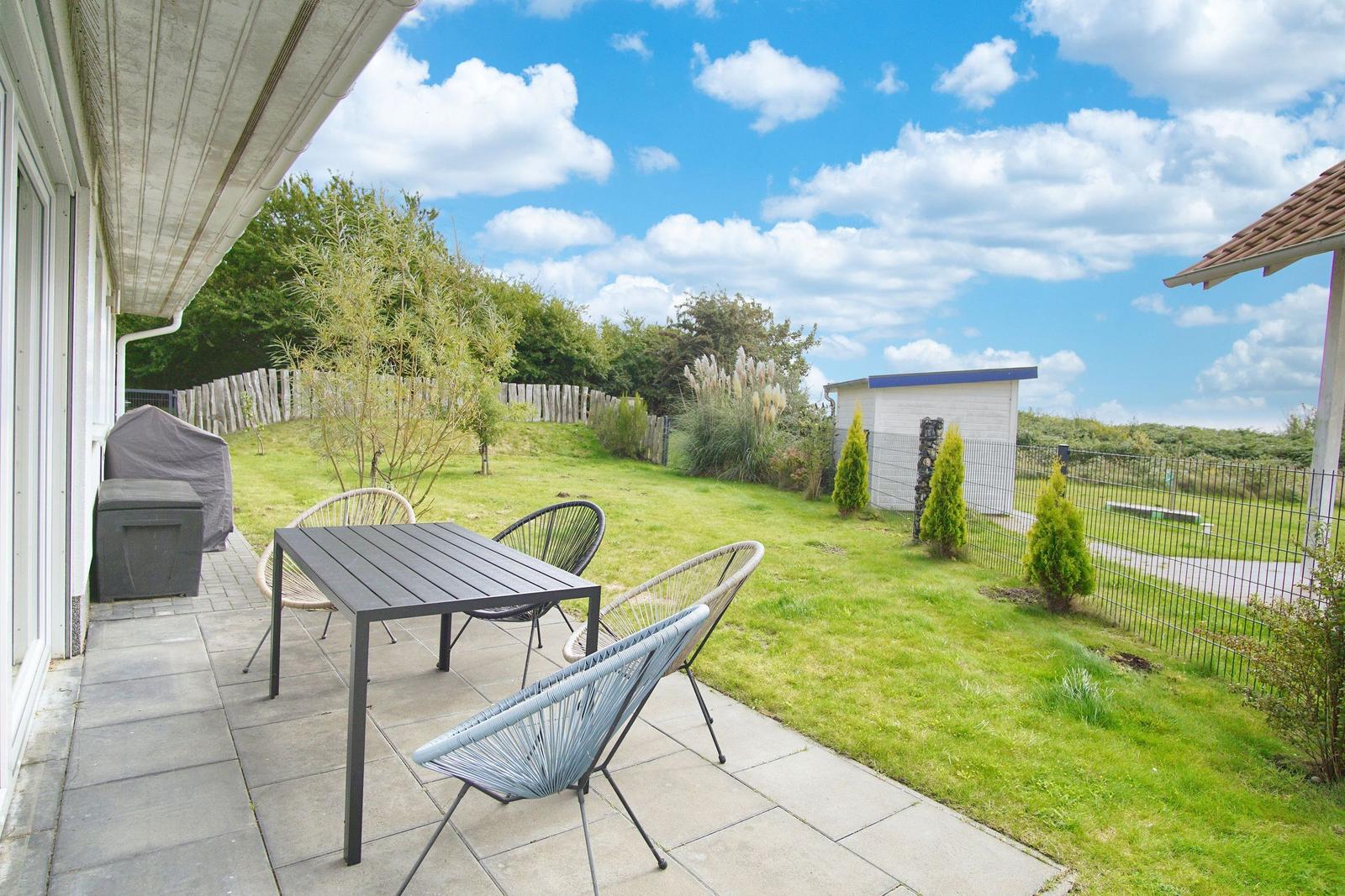 Terrasse mit Tisch und Stühlen, Grünfläche und kleinem Gartenhaus im Hintergrund.