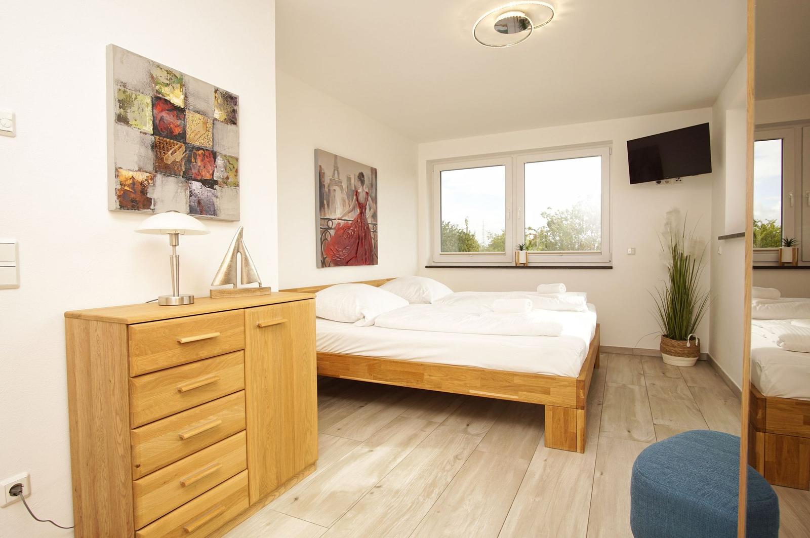 Schlafzimmer mit Holzbett, Schrank und Fenster. Wandbilder und Pflanze sichtbar.