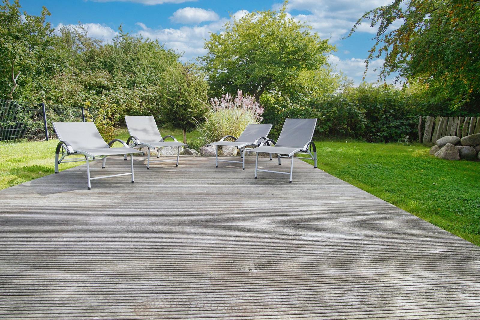 Vier Liegestühle auf einer Holzterrasse im Garten mit Gras und Bäumen.