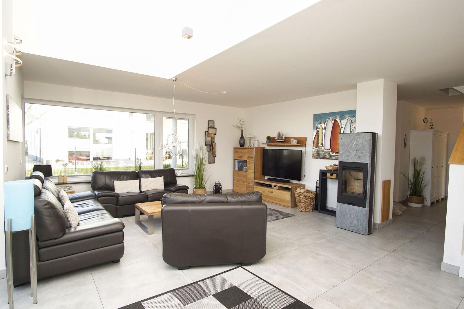 Wohnzimmer mit Ledersofa, Fernseher und Kaminofen. Große Fenster und helles Ambiente.