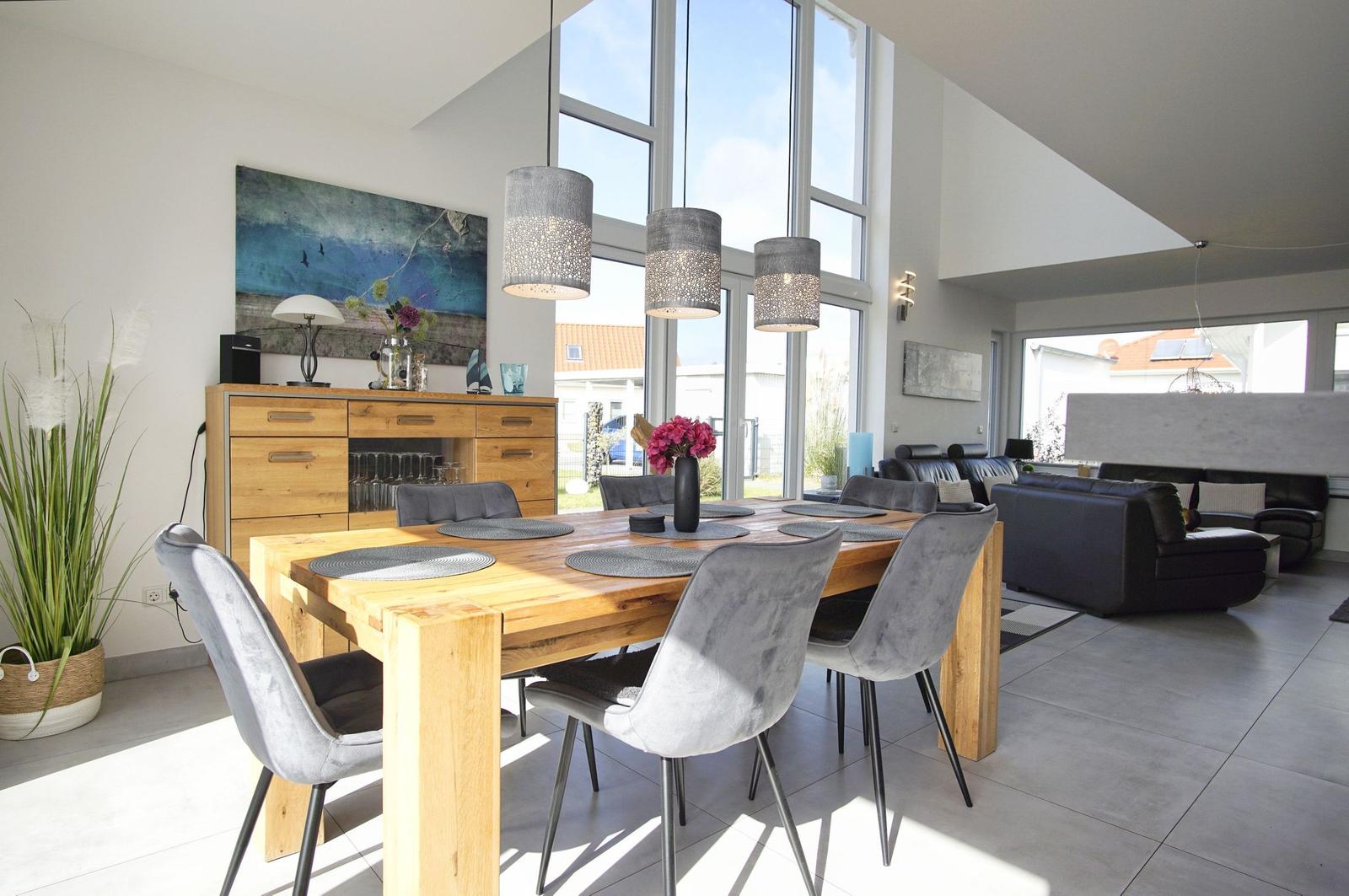 Essbereich mit Holztisch und grauen Stühlen. Große Fenster und Wohnbereich im Hintergrund.