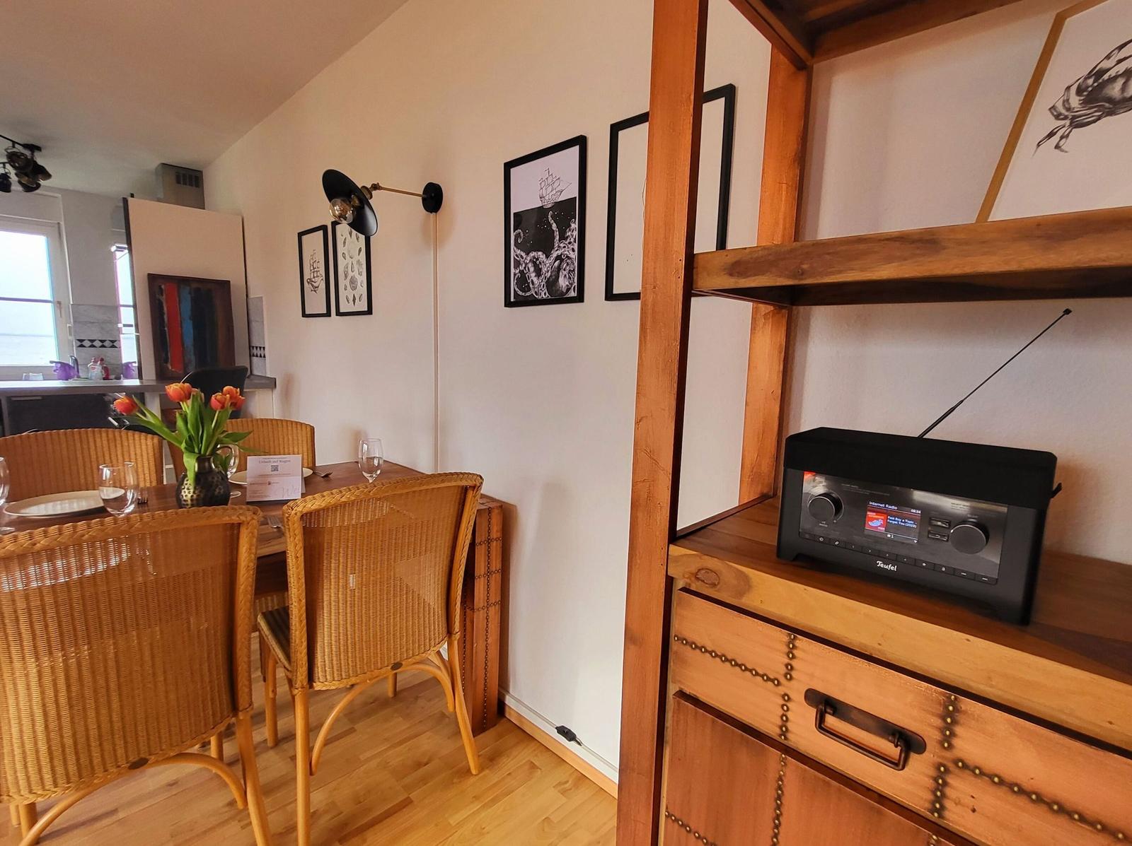 Essbereich mit Holztisch, Stühlen und Wandbildern. Radio auf Holzregal.