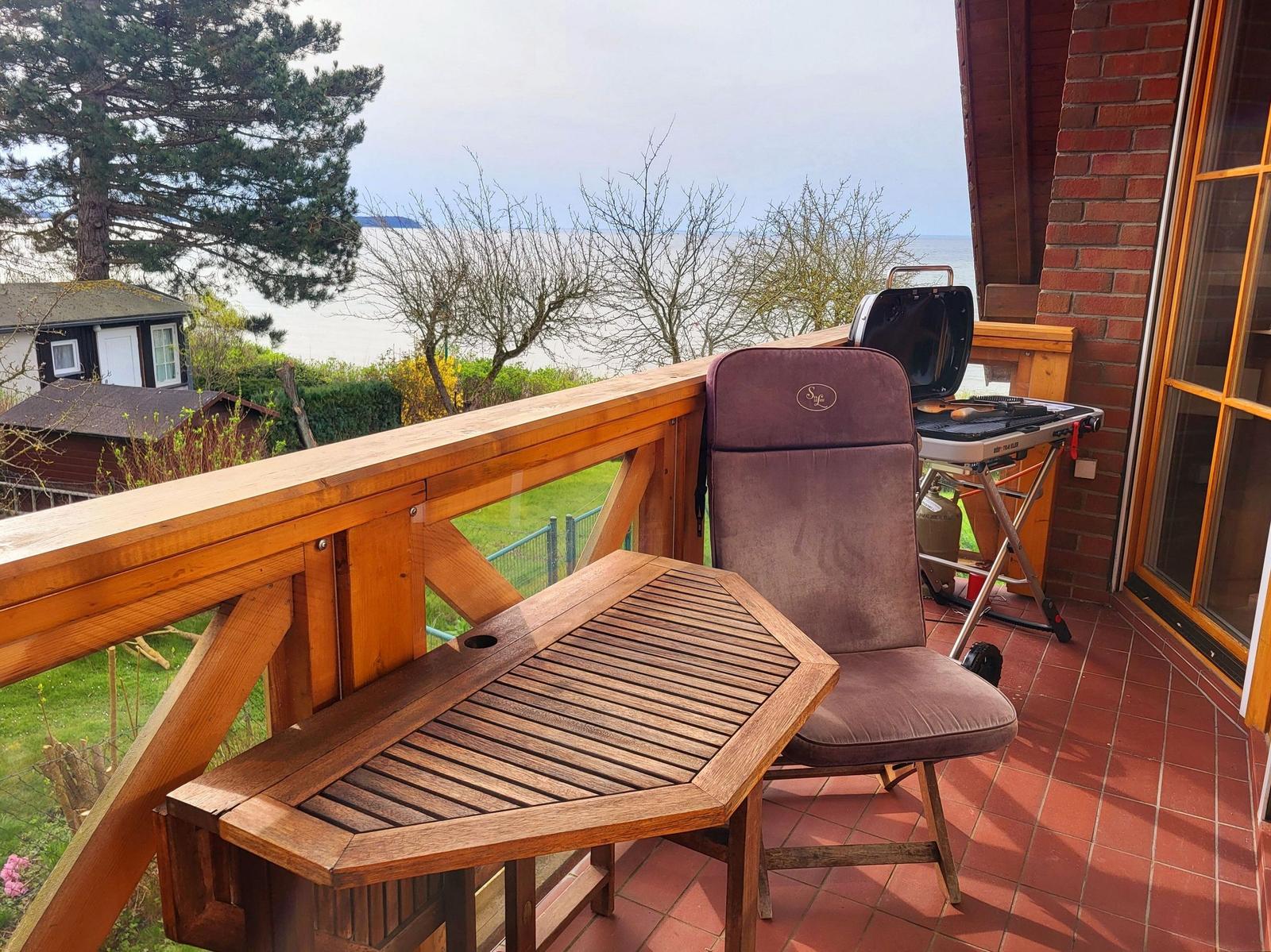 Terrasse mit Holztisch, Stuhl und Grill, Blick auf Wasser und Bäume.