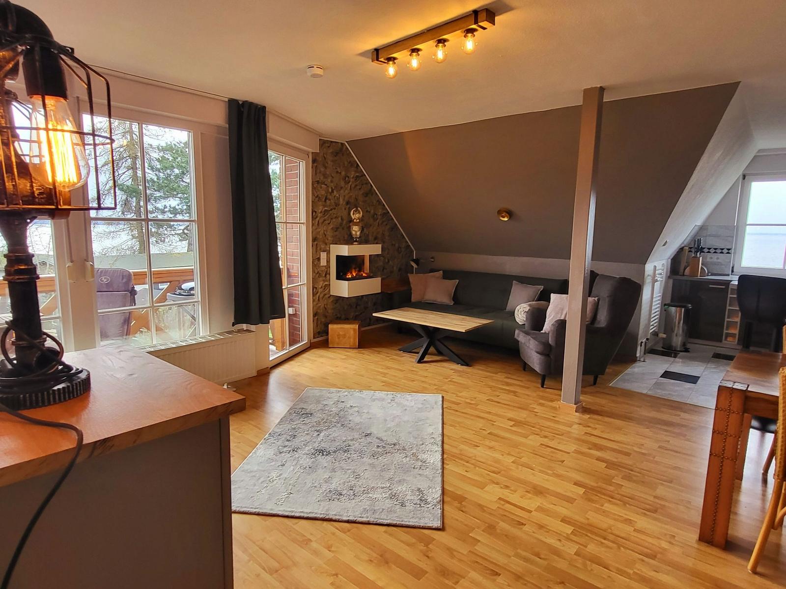 Wohnbereich mit Sofa, Sessel, Kamin und Holzboden. Küche und Terrasse sind sichtbar.