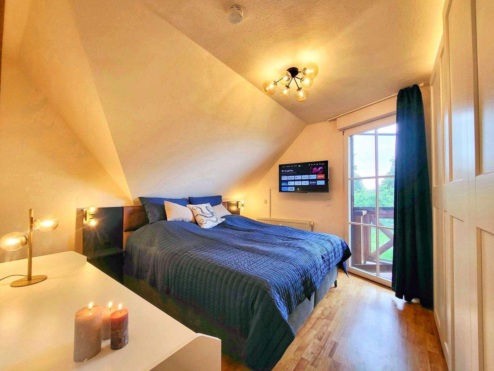Schlafzimmer mit Dachschrägen, Bett, TV und Balkontür.