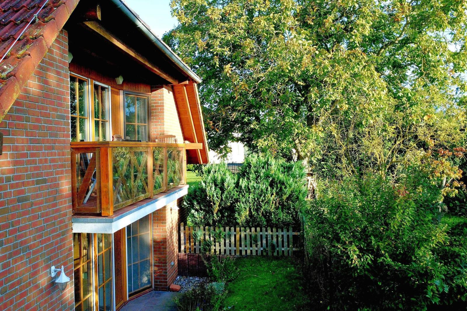 Haus mit Balkon, Garten und Bäumen. Holzgeländer und Ziegelwand sichtbar.