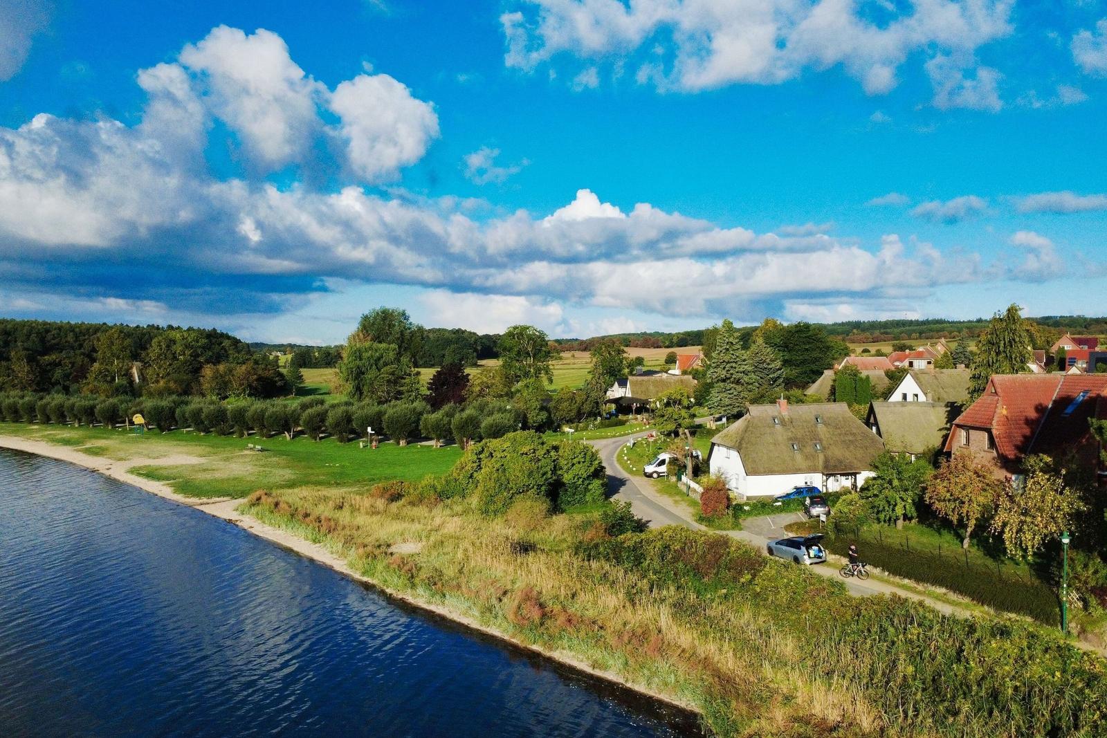 Haus am Fluss mit grünen Wiesen und Wald im Hintergrund unter blauem Himmel.