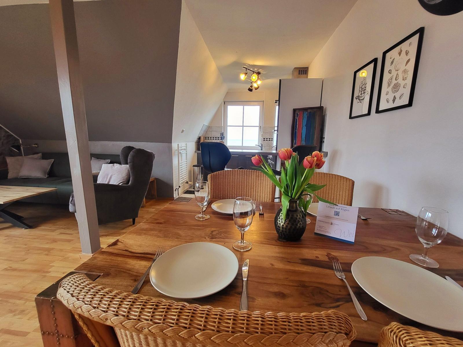 Holztisch mit Stühlen, Blumen und Geschirr. Im Hintergrund Küche und Wohnbereich.