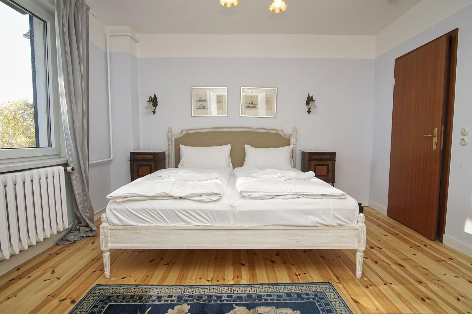 Schlafzimmer mit Doppelbett, Holzboden und Fenster. Zwei Nachttische und Wandbilder.