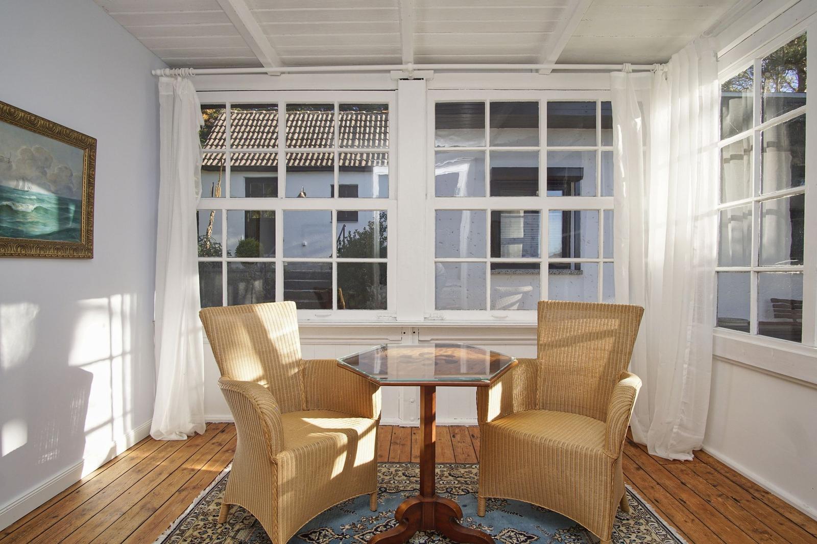 Zwei Stühle und ein Tisch stehen vor großen Fenstern mit weißen Vorhängen.