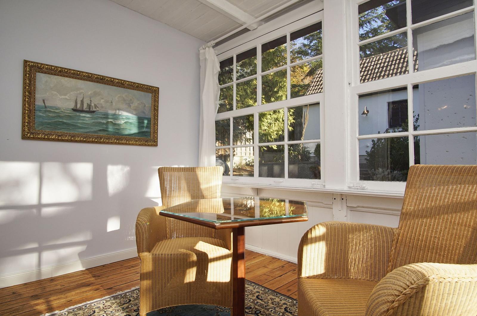 Zwei Stühle und ein Tisch stehen vor einem großen Fenster. An der Wand hängt ein Bild mit Schiffen.