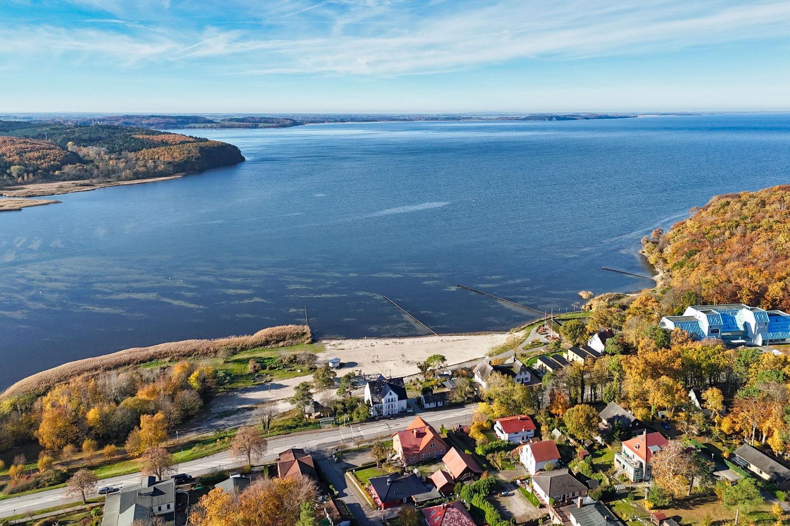 Aerial view of a coastal village with houses, beach, and large water body surrounded by autumn-colored forests.