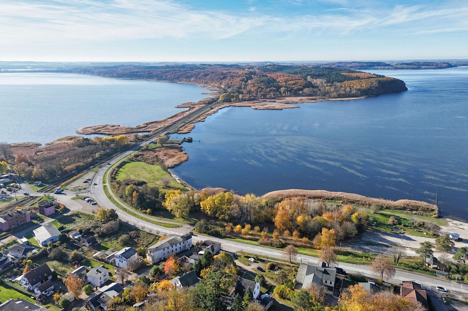Aerial view of lakeside area with road, houses, and autumn-colored trees.
