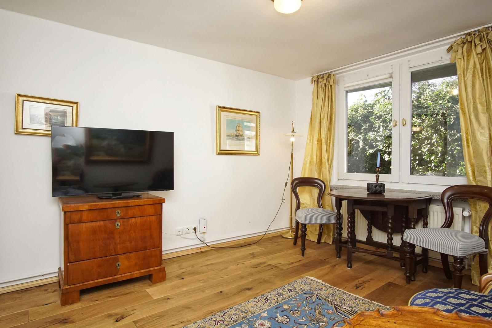 Zimmer mit Holzboden, TV auf Kommode, Tisch mit zwei Stühlen und Fenster mit Vorhängen.