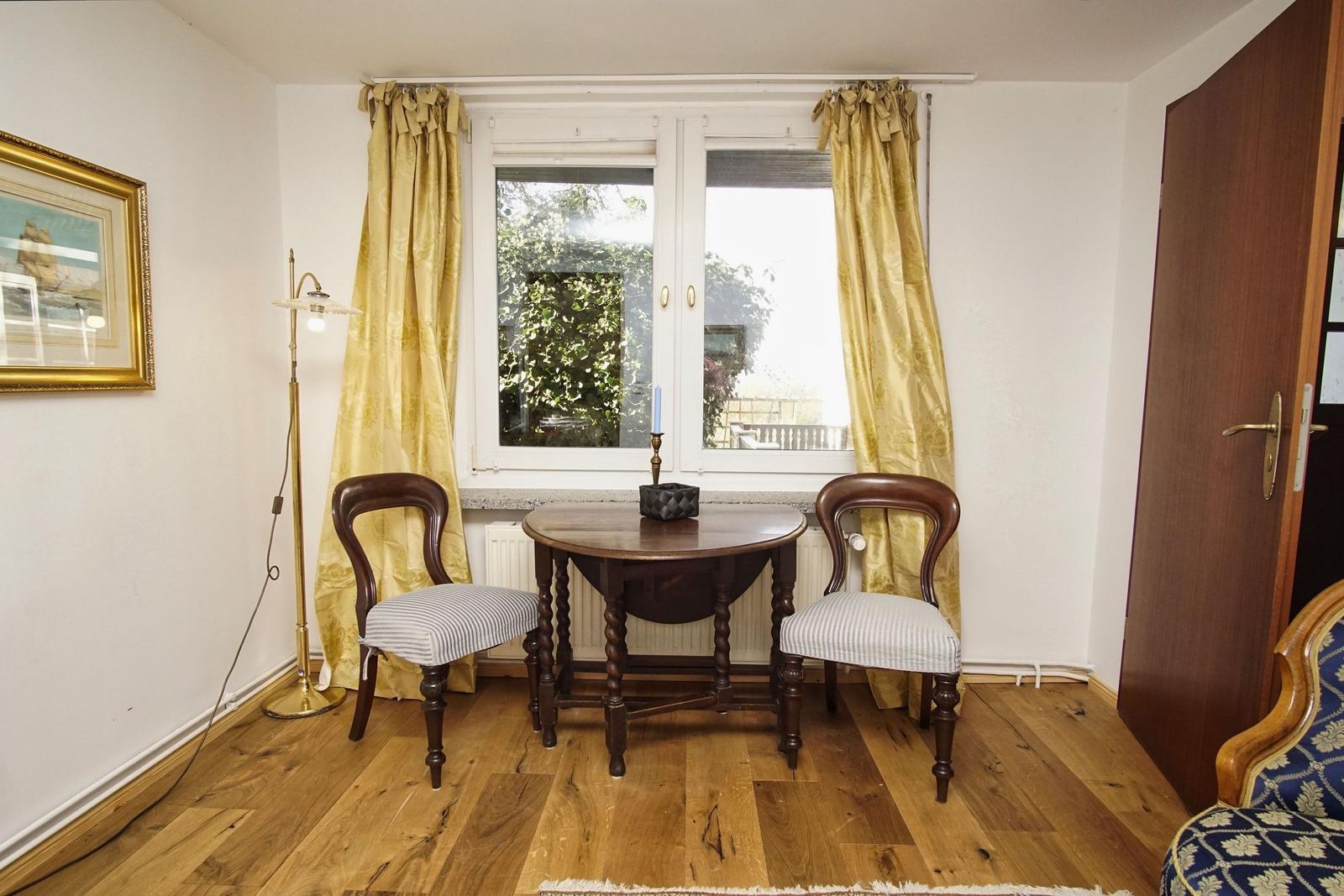 Zimmer mit Holzboden, Tisch, zwei Stühle und Fenster mit Vorhängen.