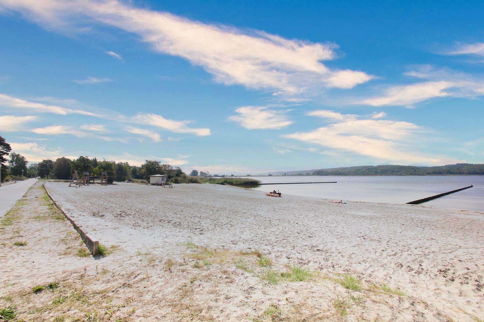 Strand mit Sand, Boot und Holzsteg am Wasser unter blauem Himmel.