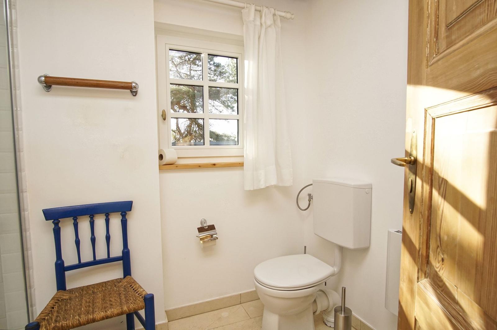 Badezimmer mit Toilette, blauer Stuhl und Fenster mit Vorhang.